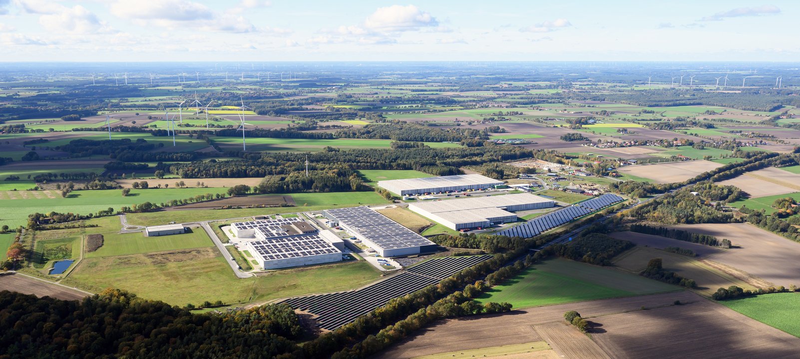 | Ⓒ Deltaland Erstellt von: Falcon Crest Air Das Bild zeigt eine Luftaufnahme mit einer weitläufigen, ländlichen Landschaft mit Feldern, Wäldern und Windkraftanlagen im Hintergrund. Im Vordergrund sind mehrere große Gebäude und Solarzellen auf Dächern zu sehen, die auf eine industrielle oder gewerbliche Nutzung hindeuten.