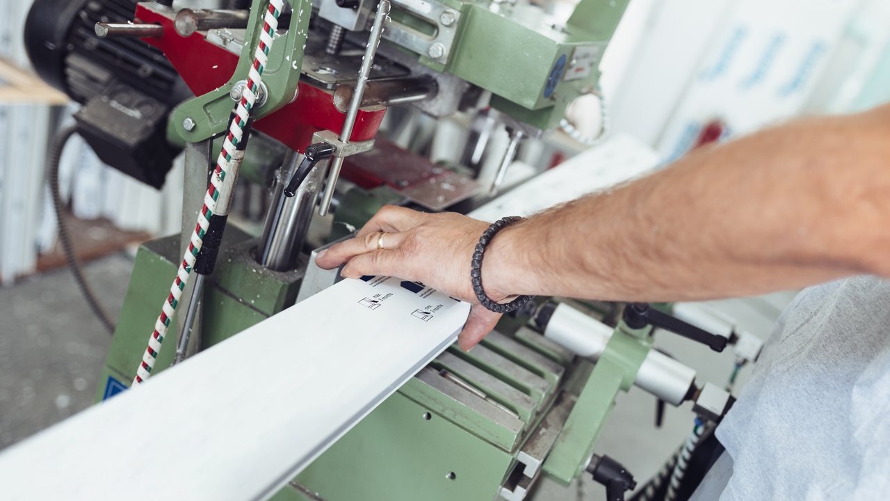  Nahaufnahme einer Hand, die ein Werkstück in eine industrielle Maschine einlegt. Die Maschine ist grün und rot, mit verschiedenen Hebeln und Schrauben. Die Szene vermittelt den Eindruck von präziser technischer Arbeit und Fertigung. 