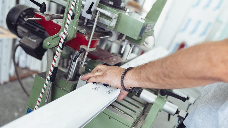 Nahaufnahme einer Hand, die ein Werkstück in eine industrielle Maschine einlegt. Die Maschine ist grün und rot, mit verschiedenen Hebeln und Schrauben. Die Szene vermittelt den Eindruck von präziser technischer Arbeit und Fertigung. 