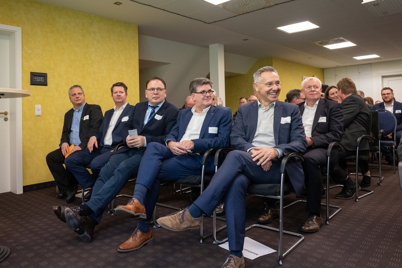 Das Bild zeigt eine Gruppe von Personen, die in der ersten Reihe einer Veranstaltung aufmerksam nach vorne blicken und offenbar einem Vortrag oder einer Präsentation folgen. Im Vordergrund sitzen fünf Personen, während im Hintergrund weitere Person zu sehen sind.