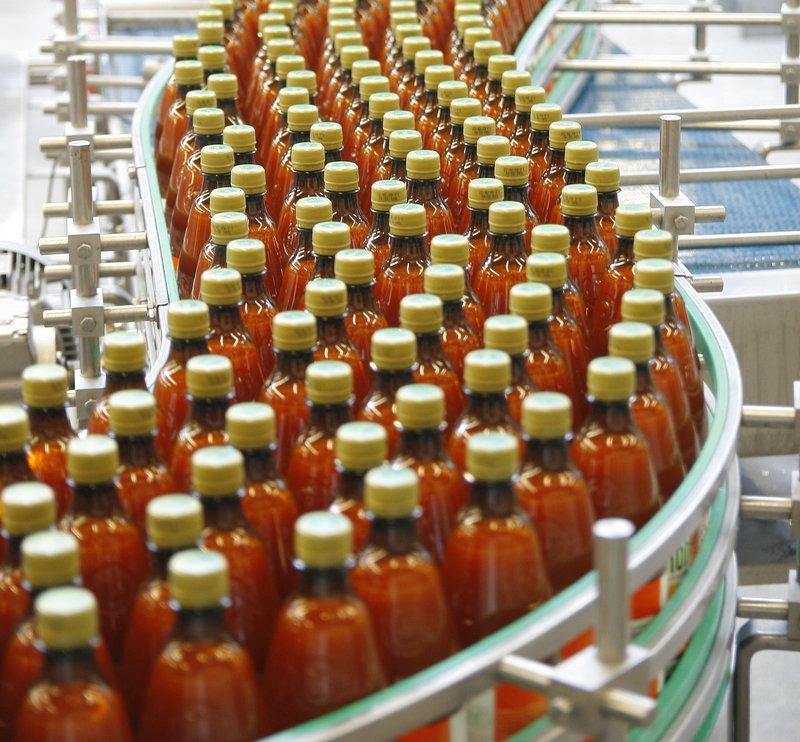 Das Bild zeigt eine Produktionslinie, auf der viele Flaschen einer roten Flüssigkeit abgefüllt und verpackt werden. Es sieht nach einem industriellen Herstellungsprozess für ein Getränk- oder Lebensmittelprodukt aus.