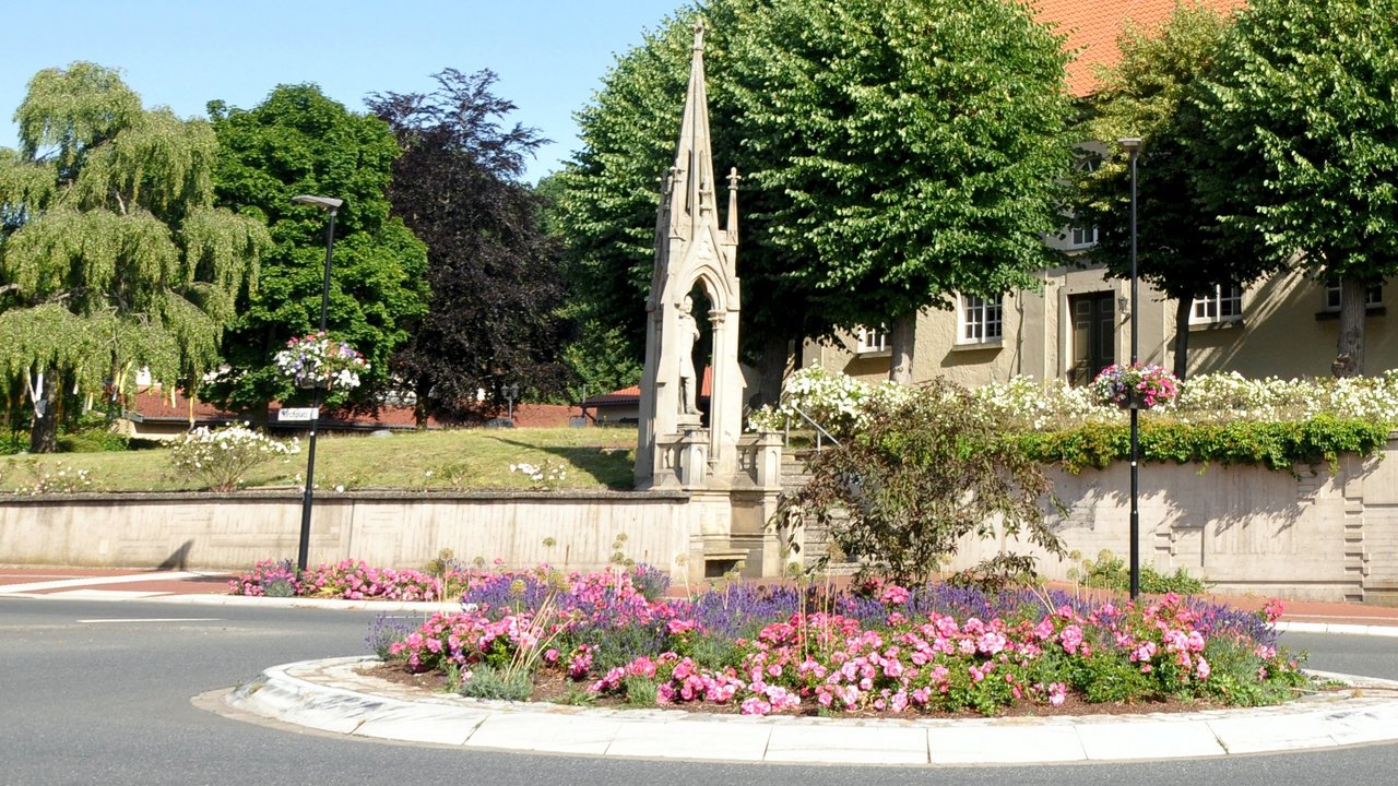  Das Bild zeigt eine malerische Kirche mit einem markanten Glockenturm, eingebettet in eine üppige, parkähnliche Umgebung. Der Kirchplatz ist mit einer Blumenrabatte und Brunnen geschmückt, was der Szene eine idyllische und einladende Atmosphäre verleiht.