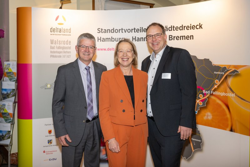 Das Bild zeigt drei Personen, die vor einem Banner mit dem Logo von DeltaLand stehen. Sie lächeln freundlich und sind in formeller Kleidung gekleidet, während sie an einer Veranstaltung teilnehmen.