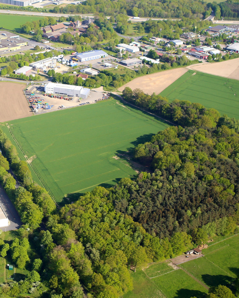 Das Bild zeigt eine Luftaufnahme einer ländlich-industriellen Gegend mit weitläufigen Feldern, einem Waldstück und mehreren Fabrik- sowie Lagergebäuden. Am oberen Bildrand sind Wohn- und Gewerbegebiete zu erkennen, die von grünen Landschaftsflächen umgeben sind.