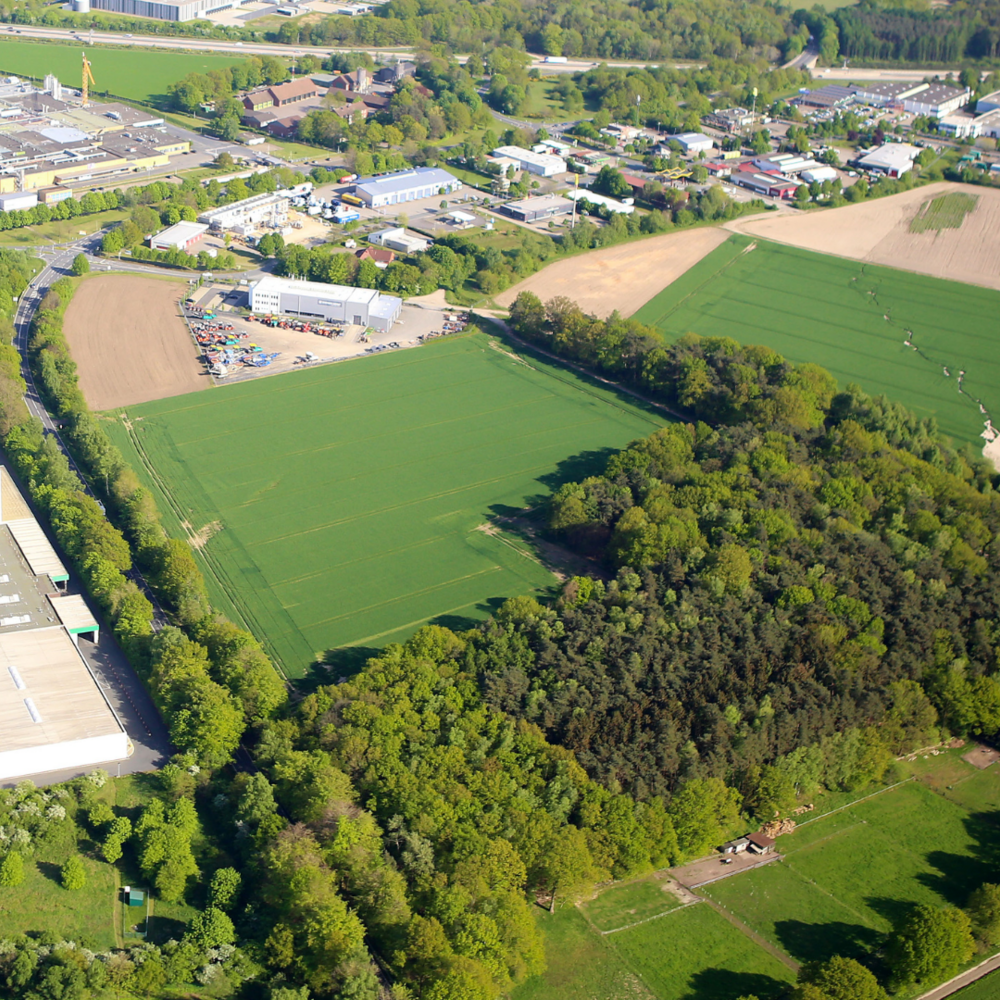 Das Bild zeigt eine Luftaufnahme einer ländlich-industriellen Gegend mit weitläufigen Feldern, einem Waldstück und mehreren Fabrik- sowie Lagergebäuden. Am oberen Bildrand sind Wohn- und Gewerbegebiete zu erkennen, die von grünen Landschaftsflächen umgeben sind.