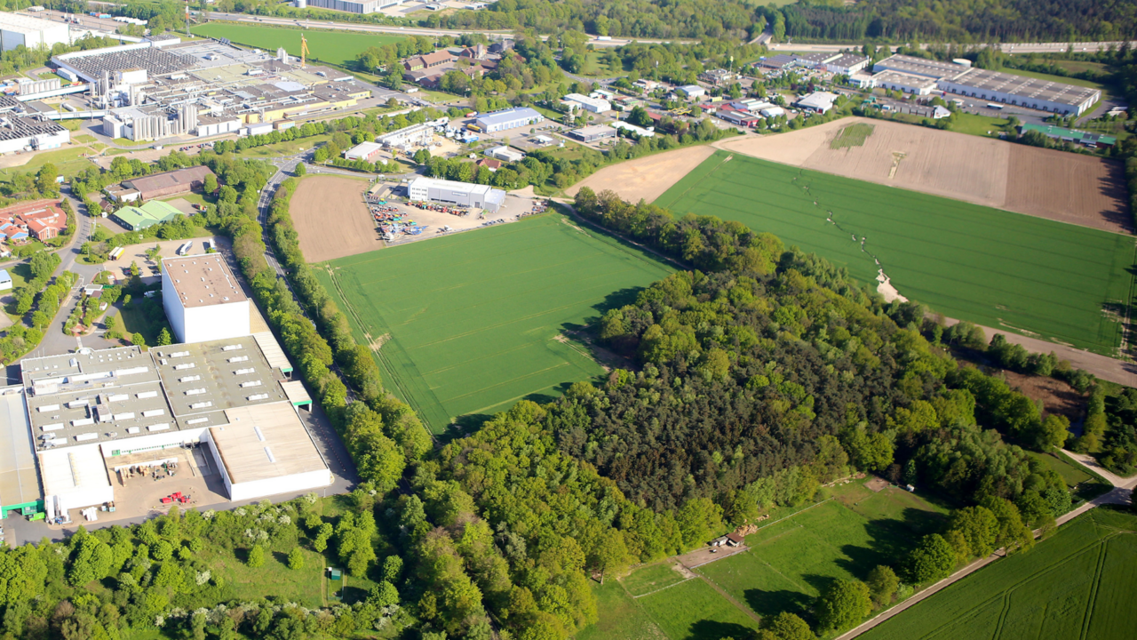 Das Bild zeigt eine Luftaufnahme einer ländlich-industriellen Gegend mit weitläufigen Feldern, einem Waldstück und mehreren Fabrik- sowie Lagergebäuden. Am oberen Bildrand sind Wohn- und Gewerbegebiete zu erkennen, die von grünen Landschaftsflächen umgeben sind.