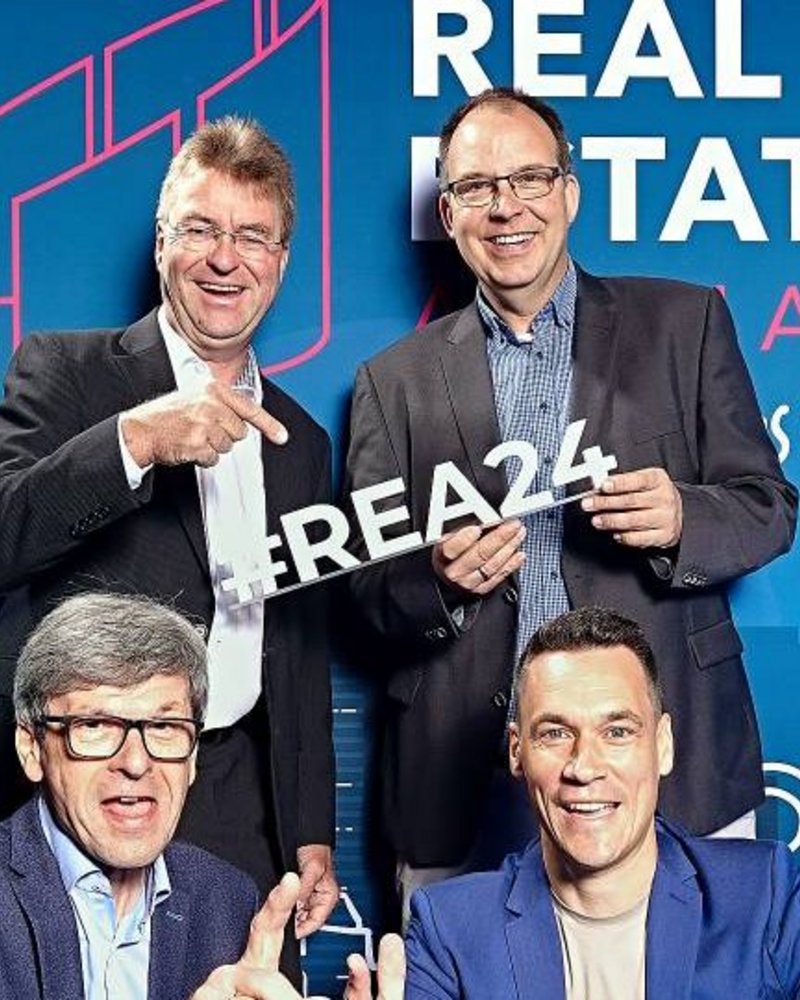 Das Bild zeigt eine Gruppe von vier Männern, die vor einer auffälligen Werbekulisse für ein Immobilienunternehmen posieren. Die Personen lachen, machen Handzeichen und scheinen die Kamera zu adressieren, was auf eine förderliche, positive Stimmung in der Gruppe hindeutet. Die Gestaltung der Kulisse in kräftigen Blau- und Rottönen sowie der Schriftzug "Real Estate" unterstreichen den werblichen Charakter der Aufnahme.