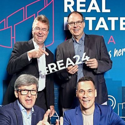 Das Bild zeigt eine Gruppe von vier Männern, die vor einer auffälligen Werbekulisse für ein Immobilienunternehmen posieren. Die Personen lachen, machen Handzeichen und scheinen die Kamera zu adressieren, was auf eine förderliche, positive Stimmung in der Gruppe hindeutet. Die Gestaltung der Kulisse in kräftigen Blau- und Rottönen sowie der Schriftzug "Real Estate" unterstreichen den werblichen Charakter der Aufnahme.