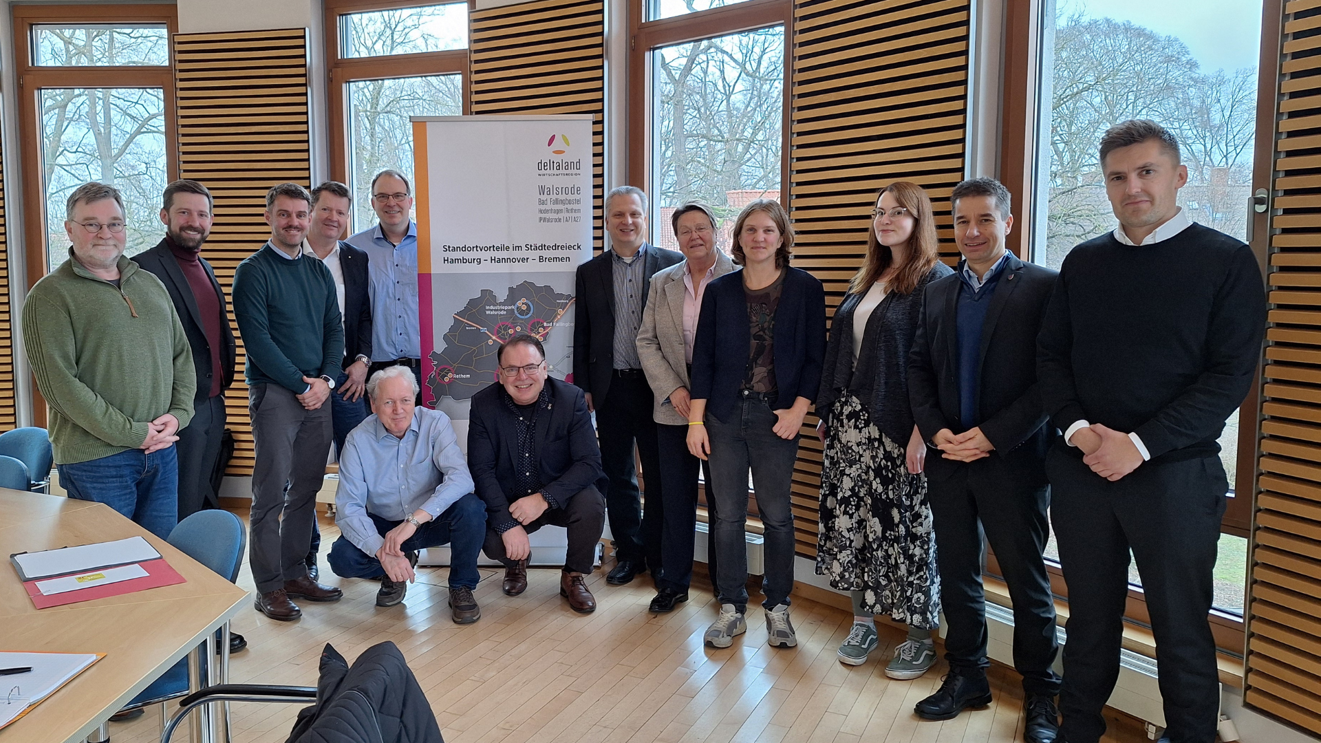 In einem hellen Raum mit großen Fenstern posiert eine Gruppe von 15 Personen für ein Gruppenfoto vor einem Banner. Die Anwesenden stehen und sitzen lächelnd zusammen.