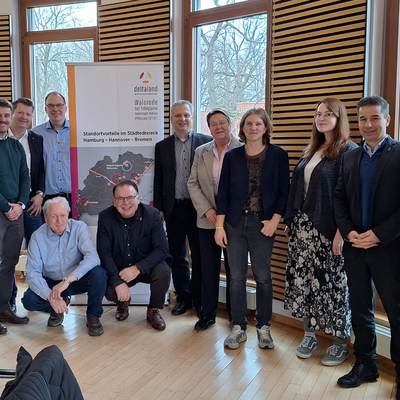 In einem hellen Raum mit großen Fenstern posiert eine Gruppe von 15 Personen für ein Gruppenfoto vor einem Banner. Die Anwesenden stehen und sitzen lächelnd zusammen.