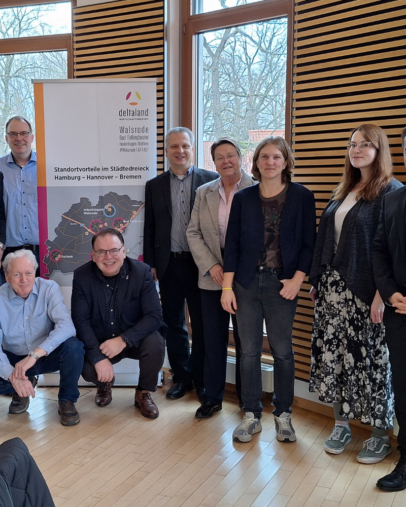 In einem hellen Raum mit großen Fenstern posiert eine Gruppe von 15 Personen für ein Gruppenfoto vor einem Banner. Die Anwesenden stehen und sitzen lächelnd zusammen.