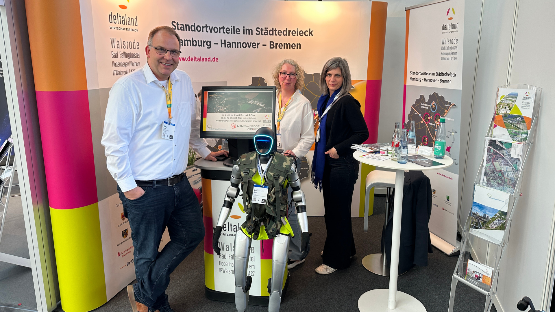 Am Deltaland-Messestand stehen ein Mann im weißen Hemd (links), eine Frau mit Brille in weißer Bluse (Mitte) und eine Frau in dunkler Jacke (rechts). Vor ihnen befindet sich ein humanoider Roboter mit Deltaland-Branding auf der Körpervorderseite. Im Hintergrund ist das Roll-up-Banner mit dem Slogan „Standortvorteile im Städtedreieck Hamburg – Hannover – Bremen" sowie ein Prospektständer mit Informationsmaterial sichtbar.