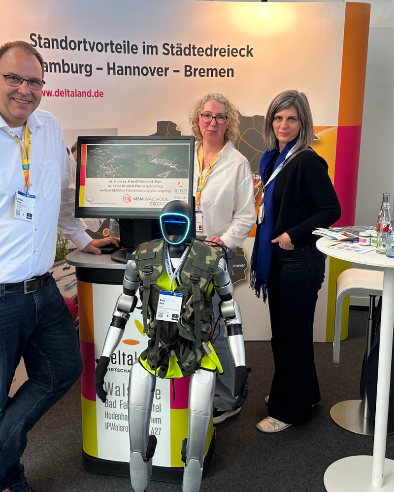 Am Deltaland-Messestand stehen ein Mann im weißen Hemd (links), eine Frau mit Brille in weißer Bluse (Mitte) und eine Frau in dunkler Jacke (rechts). Vor ihnen befindet sich ein humanoider Roboter mit Deltaland-Branding auf der Körpervorderseite. Im Hintergrund ist das Roll-up-Banner mit dem Slogan „Standortvorteile im Städtedreieck Hamburg – Hannover – Bremen" sowie ein Prospektständer mit Informationsmaterial sichtbar.