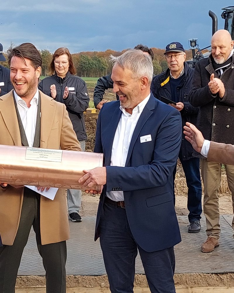 Das Bild zeigt eine Gruppe von Männern, die bei einer Grundsteinlegung eine kupferne Zeitkapsel halten. Im Hintergrund sind Zuschauer und ein Bagger zu sehen, während die Männer vorne lächeln und sich auf die Zeremonie konzentrieren.
