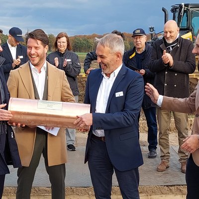 Das Bild zeigt eine Gruppe von Männern, die bei einer Grundsteinlegung eine kupferne Zeitkapsel halten. Im Hintergrund sind Zuschauer und ein Bagger zu sehen, während die Männer vorne lächeln und sich auf die Zeremonie konzentrieren.