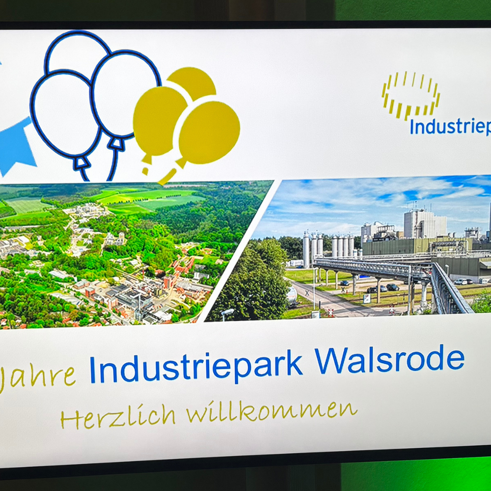 Bildschirm mit der Willkommensfolie „25 Jahre Industriepark Walsrode – Herzlich willkommen”. Die Folie zeigt das Logo des Industrieparks, Luftaufnahmen des Geländes sowie Wimpelketten und Luftballons in Blau und Gelb.