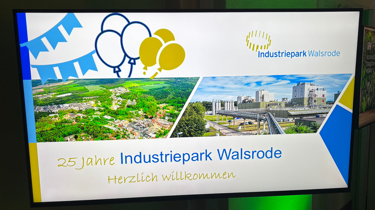 | Ⓒ Deltaland Bildschirm mit der Willkommensfolie „25 Jahre Industriepark Walsrode – Herzlich willkommen”. Die Folie zeigt das Logo des Industrieparks, Luftaufnahmen des Geländes sowie Wimpelketten und Luftballons in Blau und Gelb.