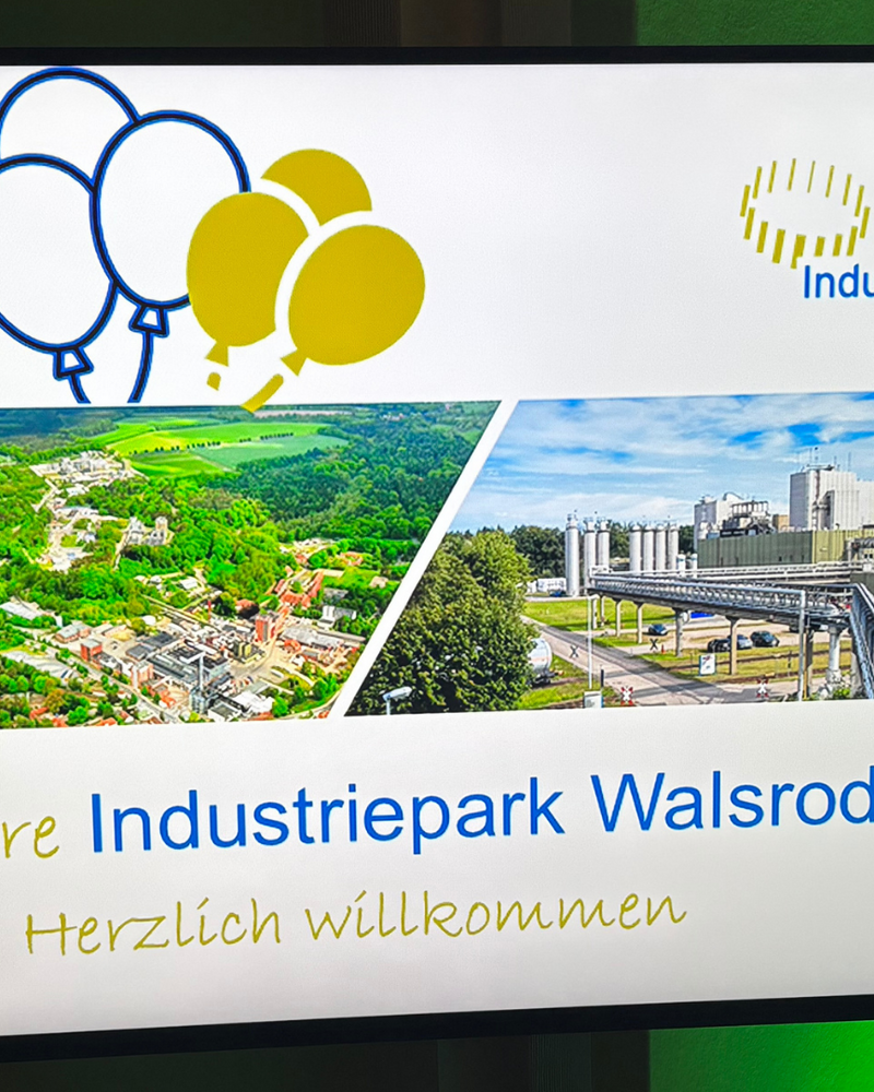 Bildschirm mit der Willkommensfolie „25 Jahre Industriepark Walsrode – Herzlich willkommen”. Die Folie zeigt das Logo des Industrieparks, Luftaufnahmen des Geländes sowie Wimpelketten und Luftballons in Blau und Gelb.