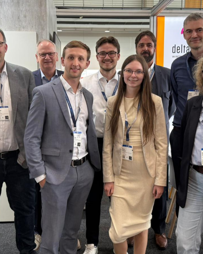 Acht Personen in Business-Kleidung stehen lächelnd für ein Gruppenfoto am deltaland-Messestand, alle tragen Besucherausweise am Lanyard. Im Hintergrund ist das deltaland-Logo auf einer beleuchteten Wand sowie eine Regionalkarte mit Standortmarkierungen zu sehen. Links eine rote Infotafel mit Grundrissplänen, rechts ein Hinweisschild mit der Standnummer „G24".
