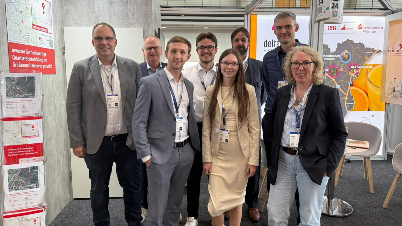 | Ⓒ Deltaland Acht Personen in Business-Kleidung stehen lächelnd für ein Gruppenfoto am deltaland-Messestand, alle tragen Besucherausweise am Lanyard. Im Hintergrund ist das deltaland-Logo auf einer beleuchteten Wand sowie eine Regionalkarte mit Standortmarkierungen zu sehen. Links eine rote Infotafel mit Grundrissplänen, rechts ein Hinweisschild mit der Standnummer „G24".
