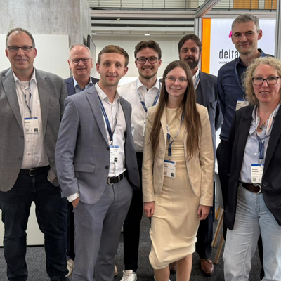 Acht Personen in Business-Kleidung stehen lächelnd für ein Gruppenfoto am deltaland-Messestand, alle tragen Besucherausweise am Lanyard. Im Hintergrund ist das deltaland-Logo auf einer beleuchteten Wand sowie eine Regionalkarte mit Standortmarkierungen zu sehen. Links eine rote Infotafel mit Grundrissplänen, rechts ein Hinweisschild mit der Standnummer „G24".