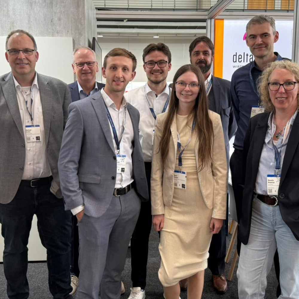 Acht Personen in Business-Kleidung stehen lächelnd für ein Gruppenfoto am deltaland-Messestand, alle tragen Besucherausweise am Lanyard. Im Hintergrund ist das deltaland-Logo auf einer beleuchteten Wand sowie eine Regionalkarte mit Standortmarkierungen zu sehen. Links eine rote Infotafel mit Grundrissplänen, rechts ein Hinweisschild mit der Standnummer „G24".