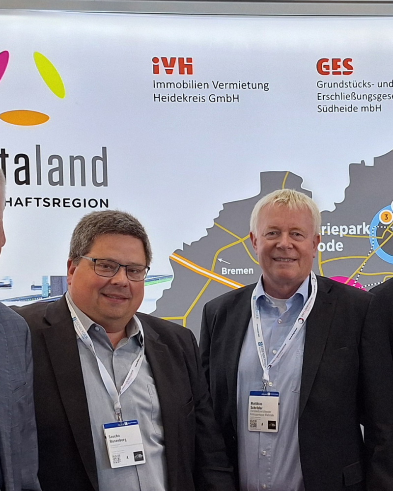 Das Bild zeigt vier Männer in Businesskleidung, die gemeinsam vor einem Messestand der Wirtschaftsregion „Deltaland“ stehen. Im Hintergrund sind Logos von regionalen Unternehmen und Institutionen wie der Kreissparkasse Walsrode sowie eine Karte mit den Standorten der Region zu sehen.