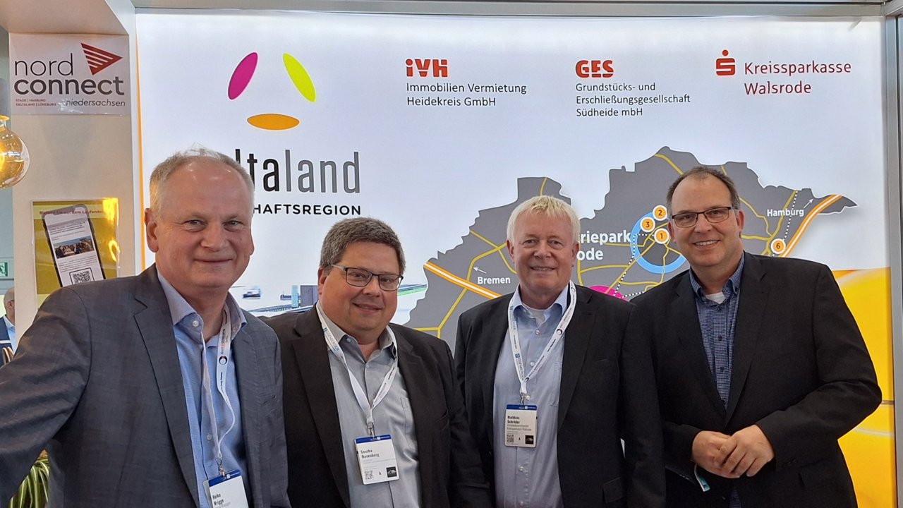 Das Bild zeigt vier Männer in Businesskleidung, die gemeinsam vor einem Messestand der Wirtschaftsregion „Deltaland“ stehen. Im Hintergrund sind Logos von regionalen Unternehmen und Institutionen wie der Kreissparkasse Walsrode sowie eine Karte mit den Standorten der Region zu sehen.