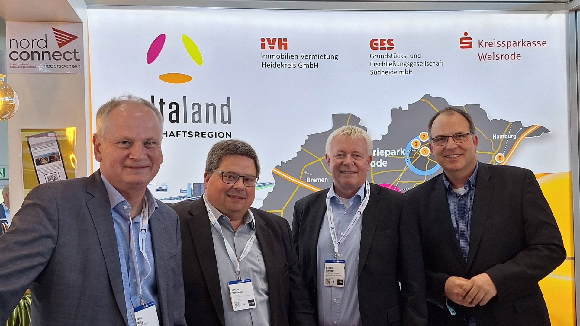 Das Bild zeigt vier Männer in Businesskleidung, die gemeinsam vor einem Messestand der Wirtschaftsregion „Deltaland“ stehen. Im Hintergrund sind Logos von regionalen Unternehmen und Institutionen wie der Kreissparkasse Walsrode sowie eine Karte mit den Standorten der Region zu sehen.