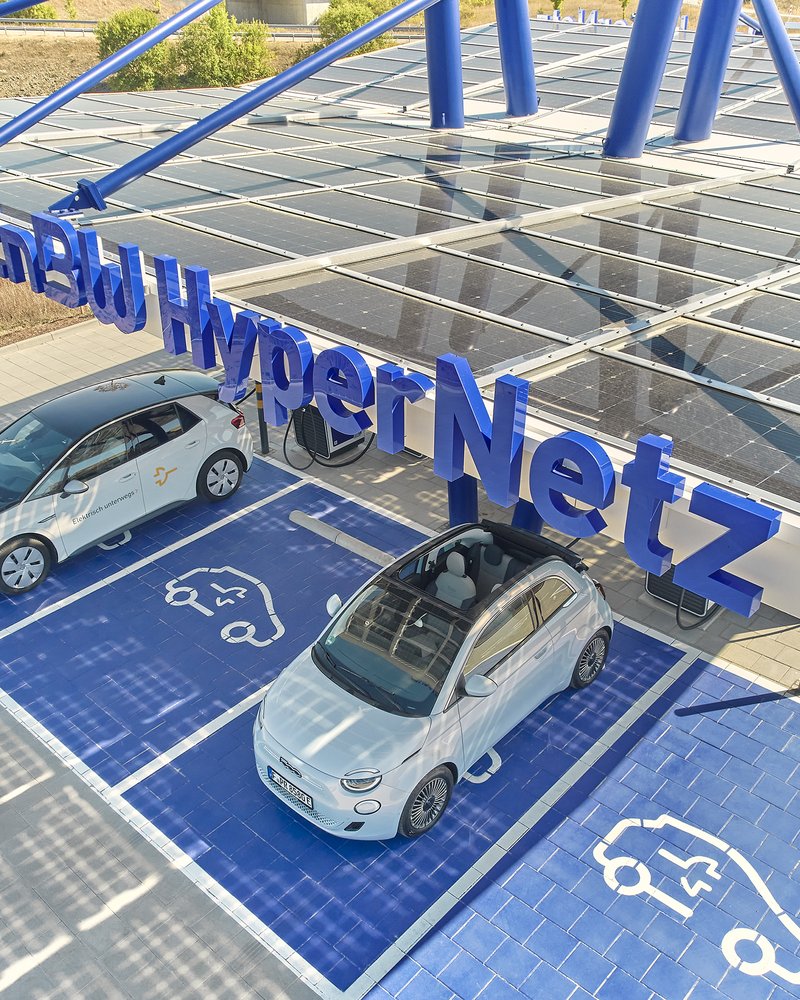 Das Bild zeigt eine große Photovoltaik-Anlage, die ein Parkplatz für Elektrofahrzeuge überdacht. Auf den Solarpaneelen sind zwei weiße Elektroautos geparkt, die offensichtlich an der Ladestation aufgeladen werden. Die Anlage vermittelt einen modernen, nachhaltigen Eindruck und bietet Autofahrern die Möglichkeit, ihre Fahrzeuge umweltfreundlich aufzuladen.