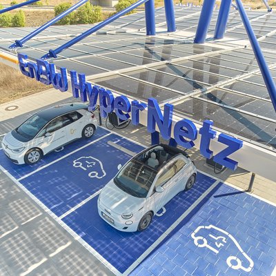 Das Bild zeigt eine große Photovoltaik-Anlage, die ein Parkplatz für Elektrofahrzeuge überdacht. Auf den Solarpaneelen sind zwei weiße Elektroautos geparkt, die offensichtlich an der Ladestation aufgeladen werden. Die Anlage vermittelt einen modernen, nachhaltigen Eindruck und bietet Autofahrern die Möglichkeit, ihre Fahrzeuge umweltfreundlich aufzuladen.