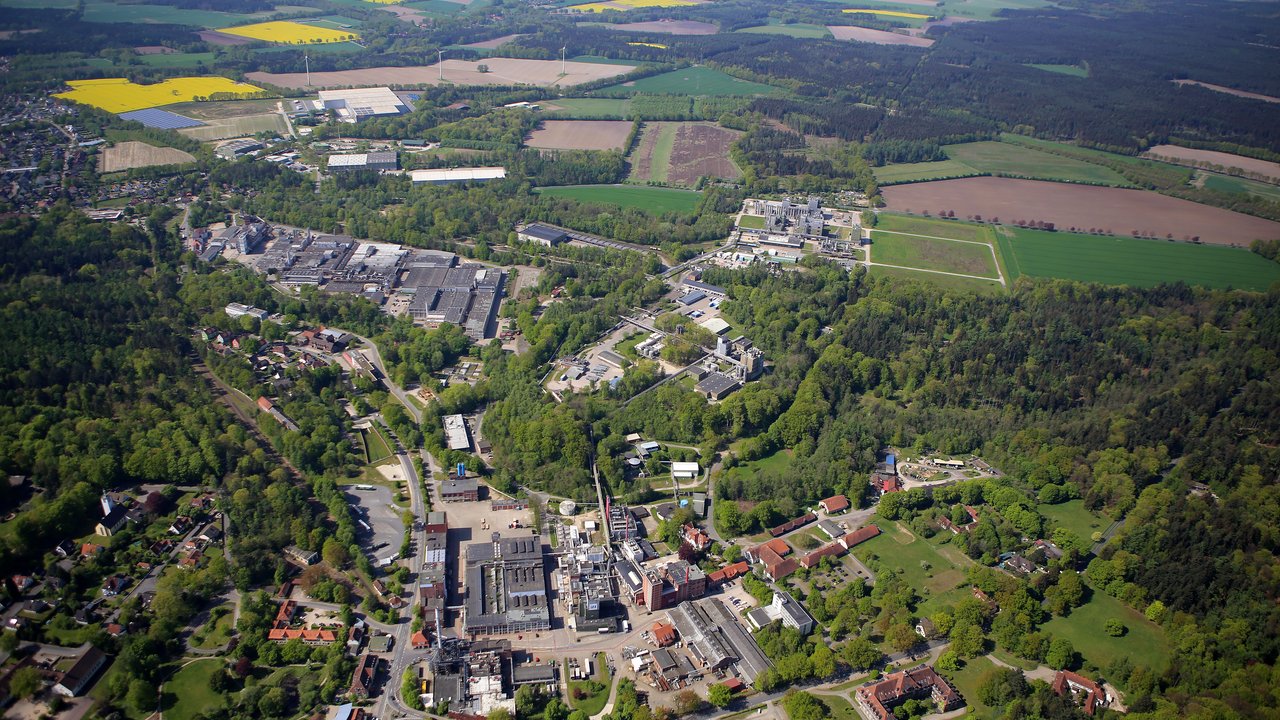 Diese Luftaufnahme zeigt eine gemischte Landschaft mit städtischer und ländlicher Prägung. Das Bild umfasst einen Industriekomplex inmitten einer grünen, ländlichen Umgebung mit Feldern, Wäldern und kleineren Siedlungen, die sich über ein weites Gebiet erstrecken.
