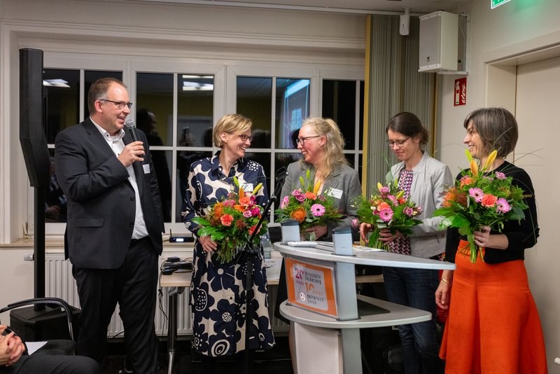 Das Bild zeigt eine Gruppe von fünf Personen, die an einem Tisch stehen und lächeln, während sie bunte Blumensträuße in den Händen halten. Die Atmosphäre wirkt festlich und feierlich, was auf eine besondere Veranstaltung oder Ehrung hinweist.