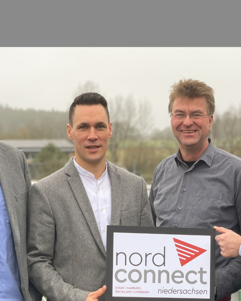 Auf dem Bild stehen vier Männer in grauen Sakkos und Hemden im Freien vor einer leicht nebligen Landschaft. Sie halten ein Schild mit dem Logo von „nord connect Niedersachsen“, das auf die Regionen Stade, Harburg, Deltaland und Lüneburg hinweist, und lächeln in die Kamera.