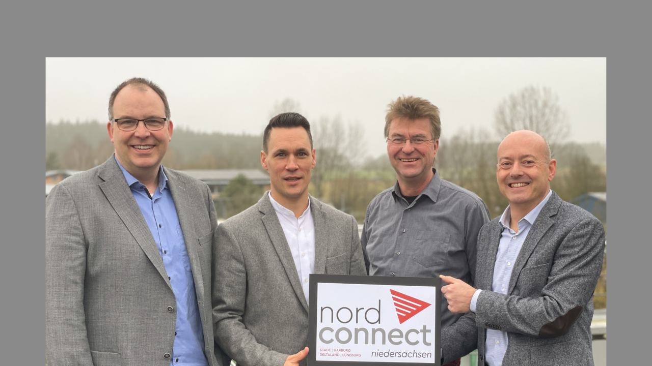 Auf dem Bild stehen vier Männer in grauen Sakkos und Hemden im Freien vor einer leicht nebligen Landschaft. Sie halten ein Schild mit dem Logo von „nord connect Niedersachsen“, das auf die Regionen Stade, Harburg, Deltaland und Lüneburg hinweist, und lächeln in die Kamera.