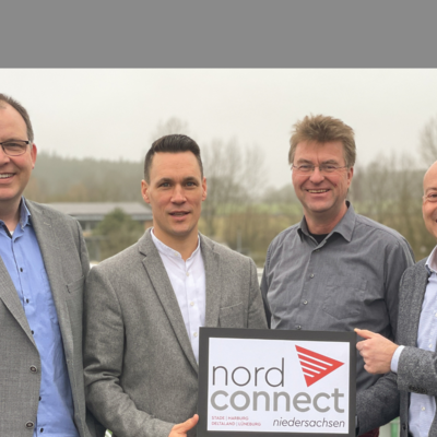 Auf dem Bild stehen vier Männer in grauen Sakkos und Hemden im Freien vor einer leicht nebligen Landschaft. Sie halten ein Schild mit dem Logo von „nord connect Niedersachsen“, das auf die Regionen Stade, Harburg, Deltaland und Lüneburg hinweist, und lächeln in die Kamera.