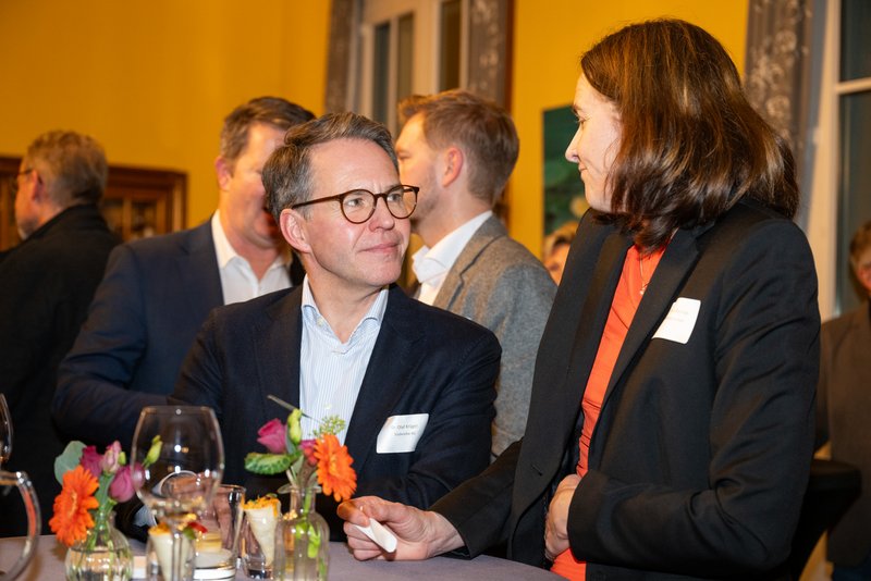 Das Bild zeigt eine gesellige Veranstaltung, bei der eine Gruppe von Menschen miteinander interagiert. Im Vordergrund lächeln ein Mann mit Brille und eine Frau, die sich freundlich unterhalten, während im Hintergrund weitere Gäste zu sehen sind.