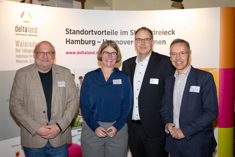 Das Bild zeigt eine Gruppe von vier Personen, die lächelnd vor einem großen Banner mit der Aufschrift "deltaland" stehen. Die Anwesenden sind in formeller Kleidung und scheinen an einer Veranstaltung teilzunehmen.