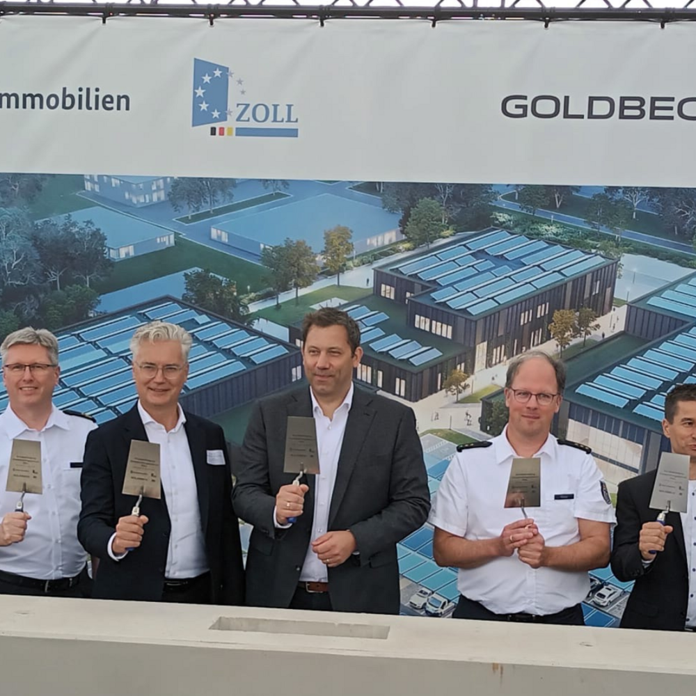Auf dem Bild stehen mehrere Personen vor einem großen Banner, die das Logo verschiedener Unternehmen zeigt, und halten symbolisch Maurerkellen in der Hand. Im Hintergrund sind Bauarbeiten und ein modernes Gebäude zu sehen, was auf den Beginn eines neuen Projekts hinweist.