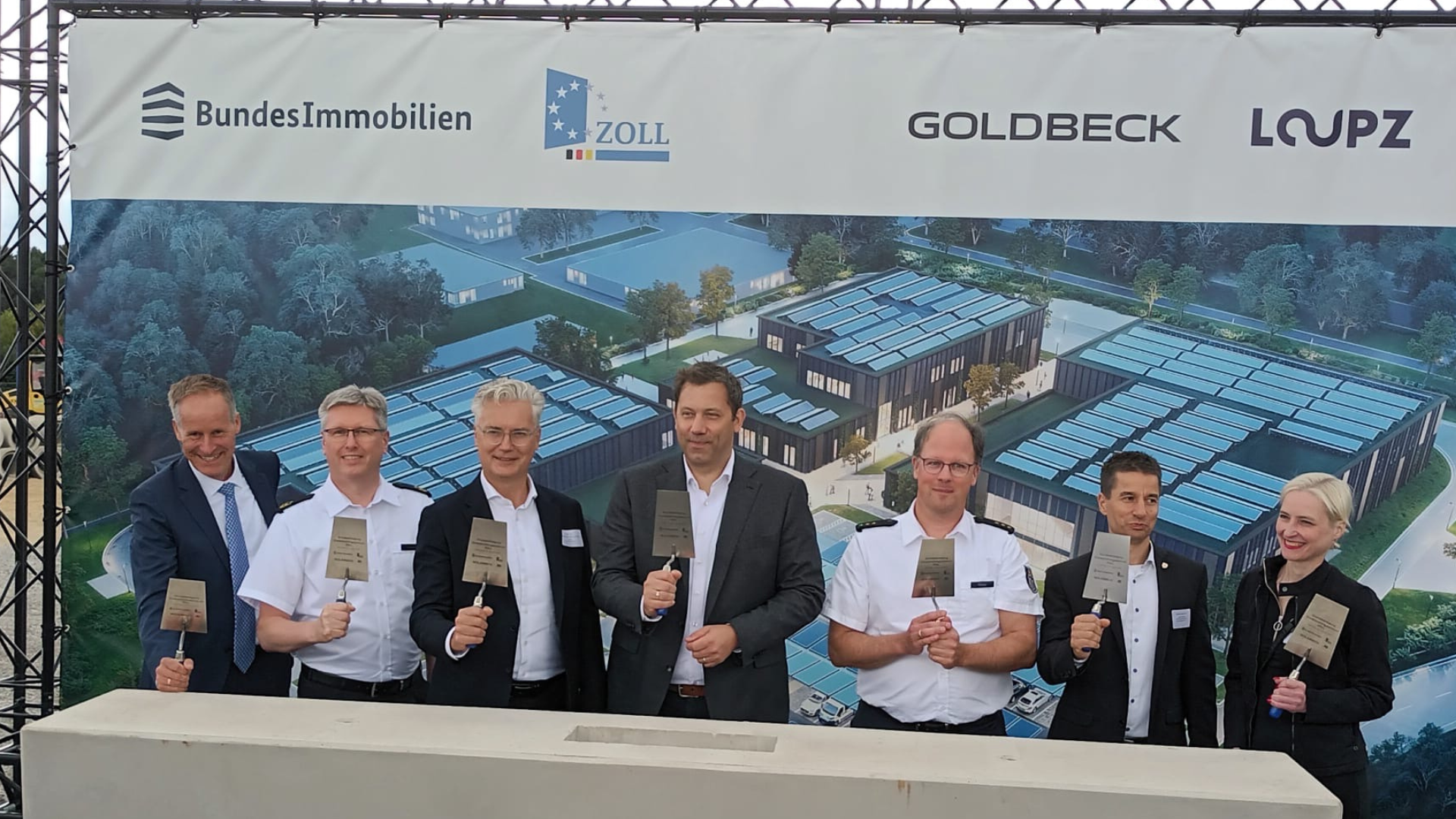 Auf dem Bild stehen mehrere Personen vor einem großen Banner, die das Logo verschiedener Unternehmen zeigt, und halten symbolisch Maurerkellen in der Hand. Im Hintergrund sind Bauarbeiten und ein modernes Gebäude zu sehen, was auf den Beginn eines neuen Projekts hinweist.