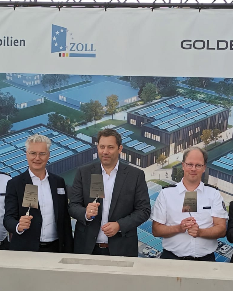 Auf dem Bild stehen mehrere Personen vor einem großen Banner, die das Logo verschiedener Unternehmen zeigt, und halten symbolisch Maurerkellen in der Hand. Im Hintergrund sind Bauarbeiten und ein modernes Gebäude zu sehen, was auf den Beginn eines neuen Projekts hinweist.