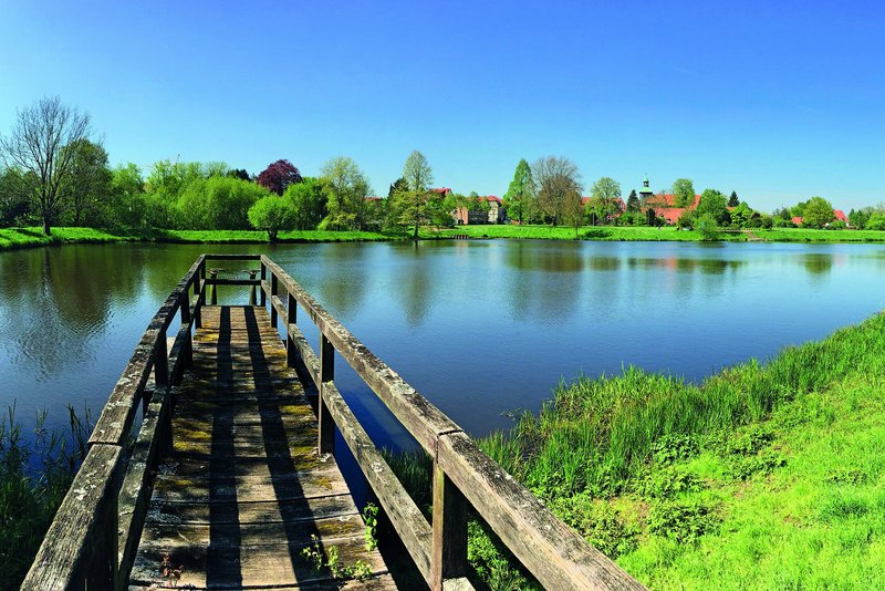 Das Bild zeigt eine idyllische Landschaft mit einem kleinen See, an dessen Ufer sich ein langer, hölzerner Steg erstreckt. Die Umgebung ist von grünen Bäumen und Wiesen gesäumt, die sich in dem ruhigen Wasser des Sees spiegeln, was eine friedliche und natürliche Atmosphäre vermittelt.