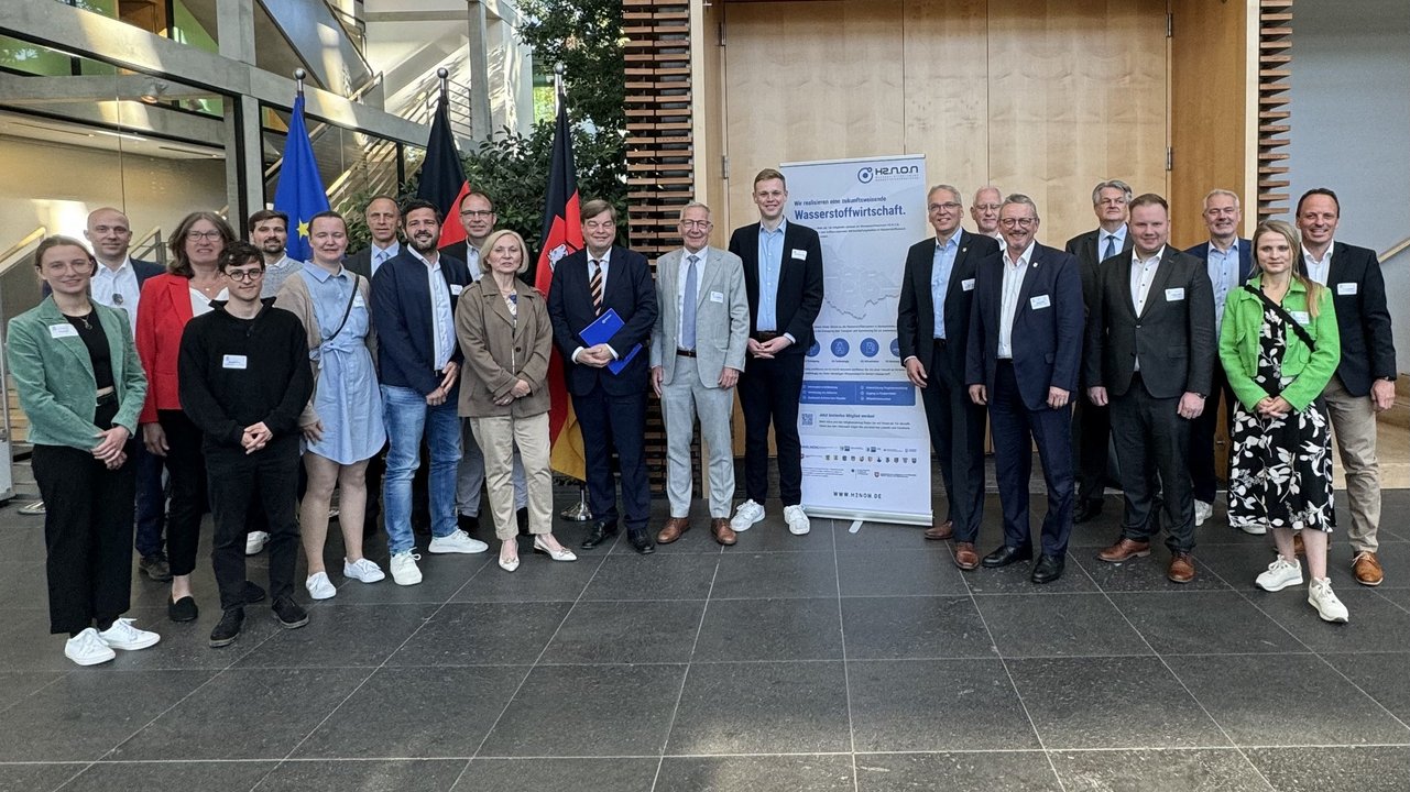Das Bild zeigt eine Gruppe von etwa zwanzig Personen in formeller Kleidung, die vor einem modernen Gebäude posieren. Im Hintergrund sind die Flaggen der Europäischen Union und Deutschlands zu sehen, sowie ein Poster mit dem Titel "Wasserstoffwirtschaft". Dies deutet auf eine Veranstaltung im Zusammenhang mit Wasserstofftechnologie hin. Die Umgebung ist modern und lichtdurchflutet, und die Teilnehmer tragen Namensschilder und halten teilweise Dokumente in der Hand. Alle blicken lächelnd in die Kamera.