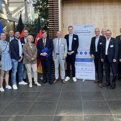 Das Bild zeigt eine Gruppe von etwa zwanzig Personen in formeller Kleidung, die vor einem modernen Gebäude posieren. Im Hintergrund sind die Flaggen der Europäischen Union und Deutschlands zu sehen, sowie ein Poster mit dem Titel "Wasserstoffwirtschaft". Dies deutet auf eine Veranstaltung im Zusammenhang mit Wasserstofftechnologie hin. Die Umgebung ist modern und lichtdurchflutet, und die Teilnehmer tragen Namensschilder und halten teilweise Dokumente in der Hand. Alle blicken lächelnd in die Kamera.