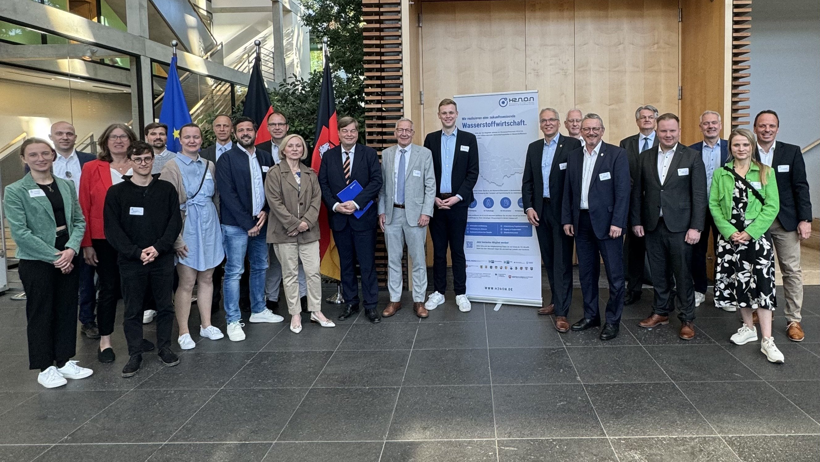 Das Bild zeigt eine Gruppe von etwa zwanzig Personen in formeller Kleidung, die vor einem modernen Gebäude posieren. Im Hintergrund sind die Flaggen der Europäischen Union und Deutschlands zu sehen, sowie ein Poster mit dem Titel "Wasserstoffwirtschaft". Dies deutet auf eine Veranstaltung im Zusammenhang mit Wasserstofftechnologie hin. Die Umgebung ist modern und lichtdurchflutet, und die Teilnehmer tragen Namensschilder und halten teilweise Dokumente in der Hand. Alle blicken lächelnd in die Kamera.