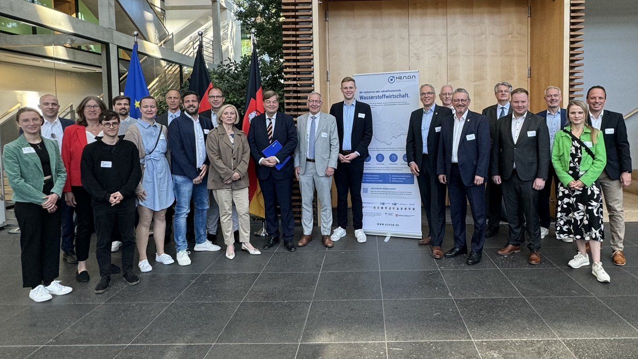 Das Bild zeigt eine Gruppe von etwa zwanzig Personen in formeller Kleidung, die vor einem modernen Gebäude posieren. Im Hintergrund sind die Flaggen der Europäischen Union und Deutschlands zu sehen, sowie ein Poster mit dem Titel "Wasserstoffwirtschaft". Dies deutet auf eine Veranstaltung im Zusammenhang mit Wasserstofftechnologie hin. Die Umgebung ist modern und lichtdurchflutet, und die Teilnehmer tragen Namensschilder und halten teilweise Dokumente in der Hand. Alle blicken lächelnd in die Kamera.