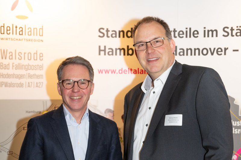 Das Bild zeigt zwei Männer, die freundlich vor einem Banner mit der Aufschrift "deltaland" stehen. Sie tragen formelle Kleidung und scheinen an einer geschäftlichen Veranstaltung teilzunehmen.