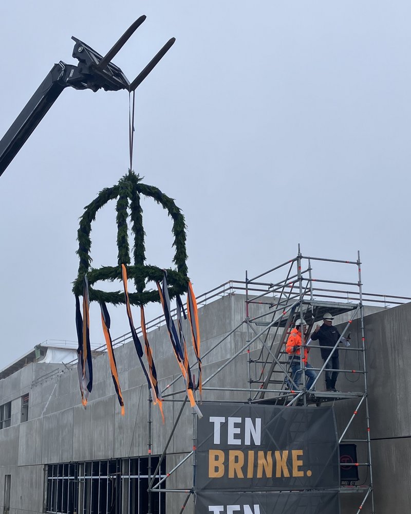 Das Bild zeigt eine Baustelle mit einem Gerüst, an dem Werbebanner für "Ten Brink" angebracht sind. Im Vordergrund befindet sich eine auffällige Skulptur oder Dekoration, die aus Stahlkonstruktionen und einem Kranz aus Grünpflanzen besteht.