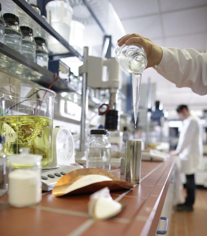 Das Bild zeigt ein Labor, in dem ein Wissenschaftler eine klare Flüssigkeit aus einem Glasbehälter gießt. Auf der Arbeitsfläche befinden sich mehrere Bechergläser mit unterschiedlichen Flüssigkeiten und Pulvern sowie Laborgeräte und Chemikalien.
