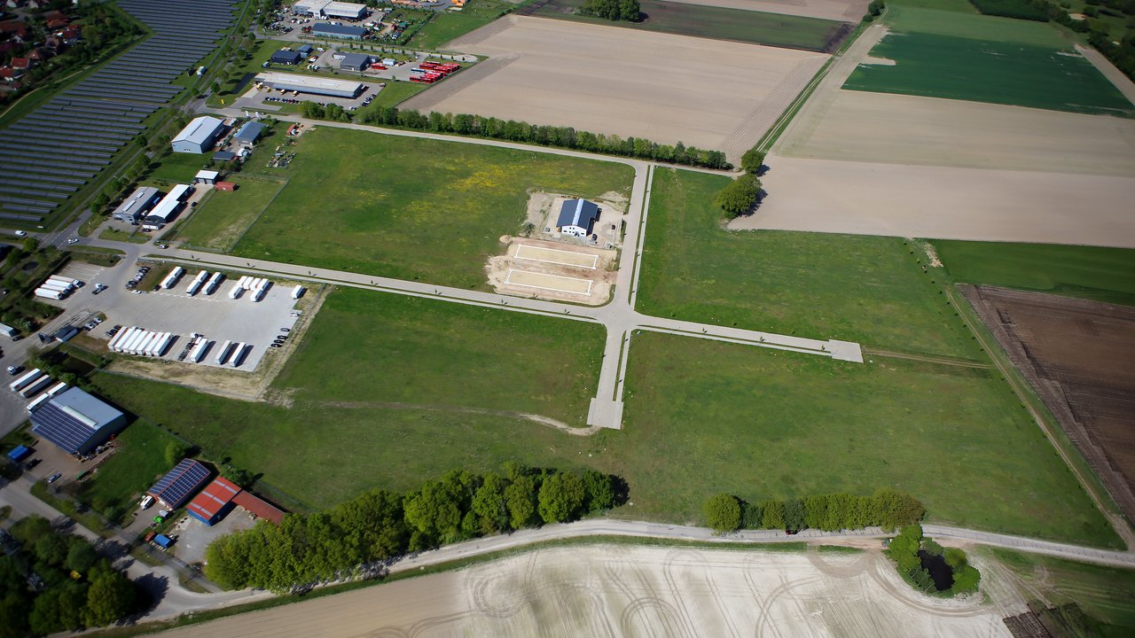  Das Bild zeigt eine ländliche Gegend mit einer Mischung aus landwirtschaftlichen Nutzflächen und industrieller Infrastruktur. Es ist eine Luftaufnahme, die einen Überblick über die Landschaft mit Feldern, Wiesen, Waldstücken und einigen Gebäuden und Lagerhallen gibt.
